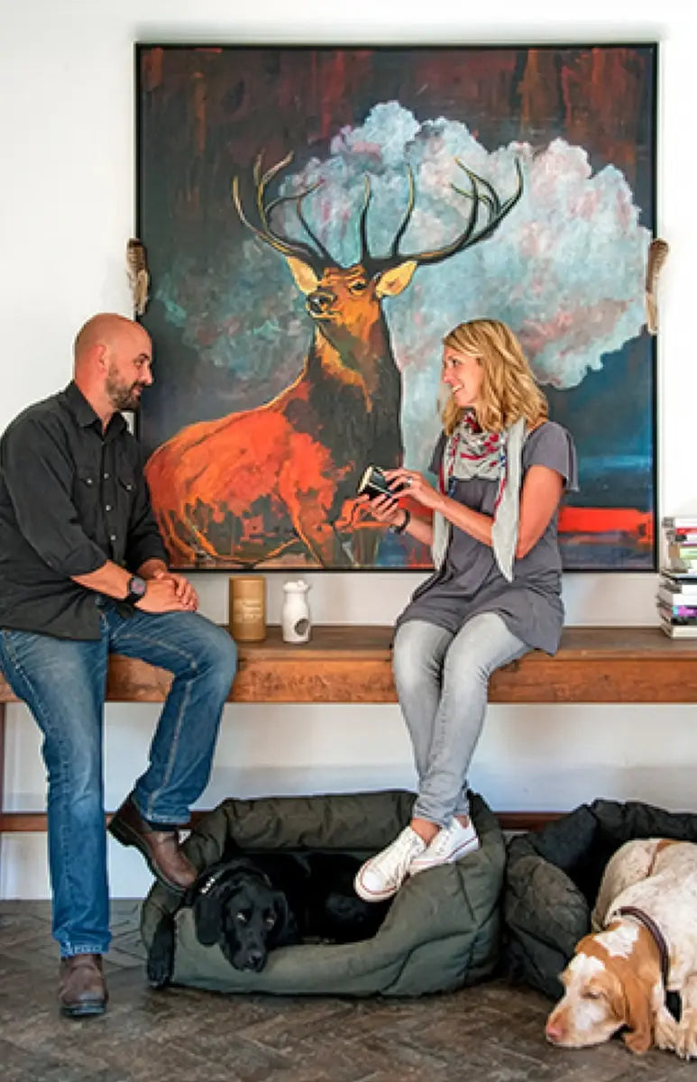 Krystyna Patey founder of Parkminster Candles sitting on a bench with the family dogs in front of a large painting of a deer. Discussing Business