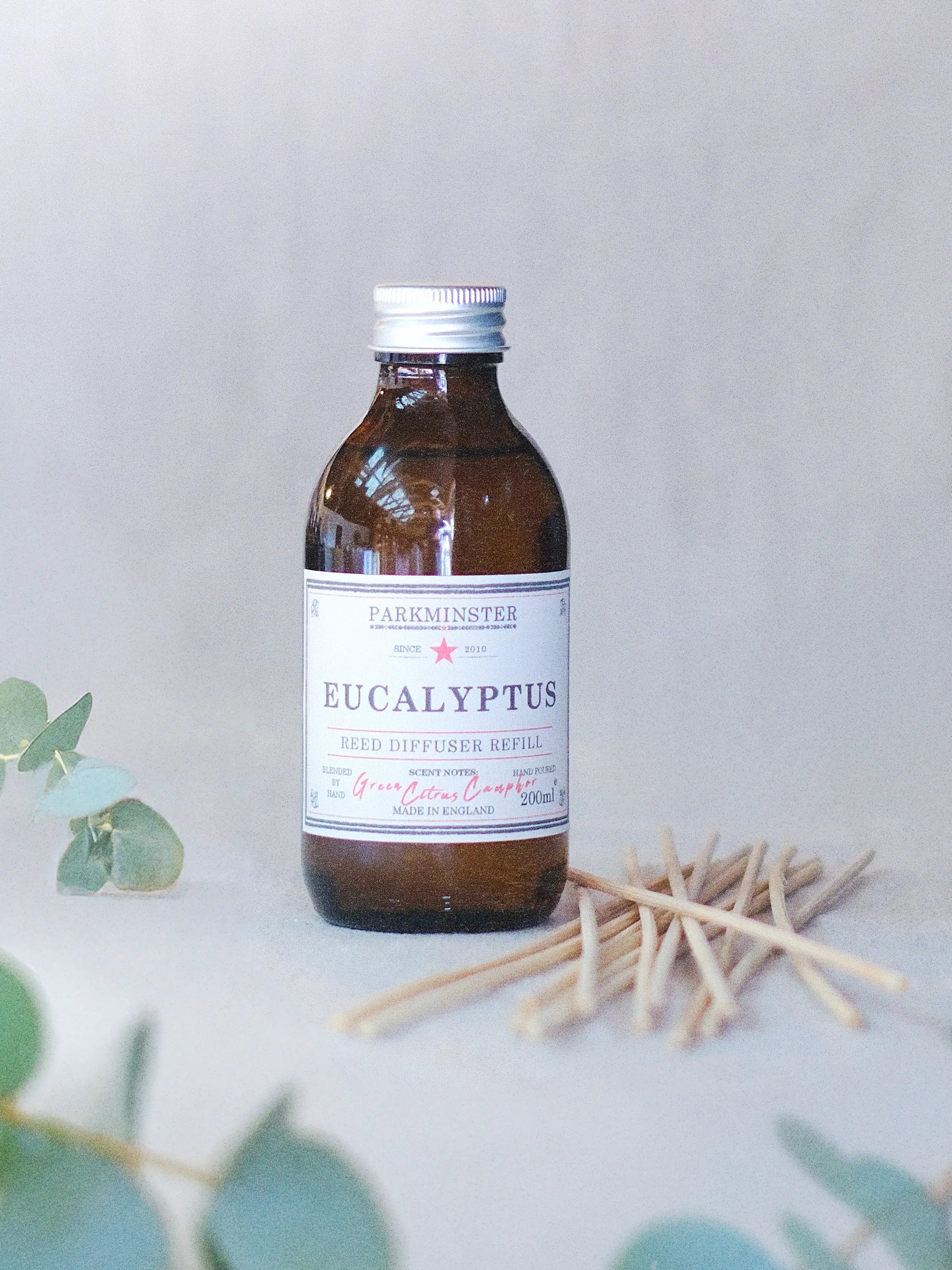 a Parkminster 100% Essential Oil Eucalyptus Reed Diffuser refill in 200ml amber glass apothecary bottle with natural rattan reeds beside it.