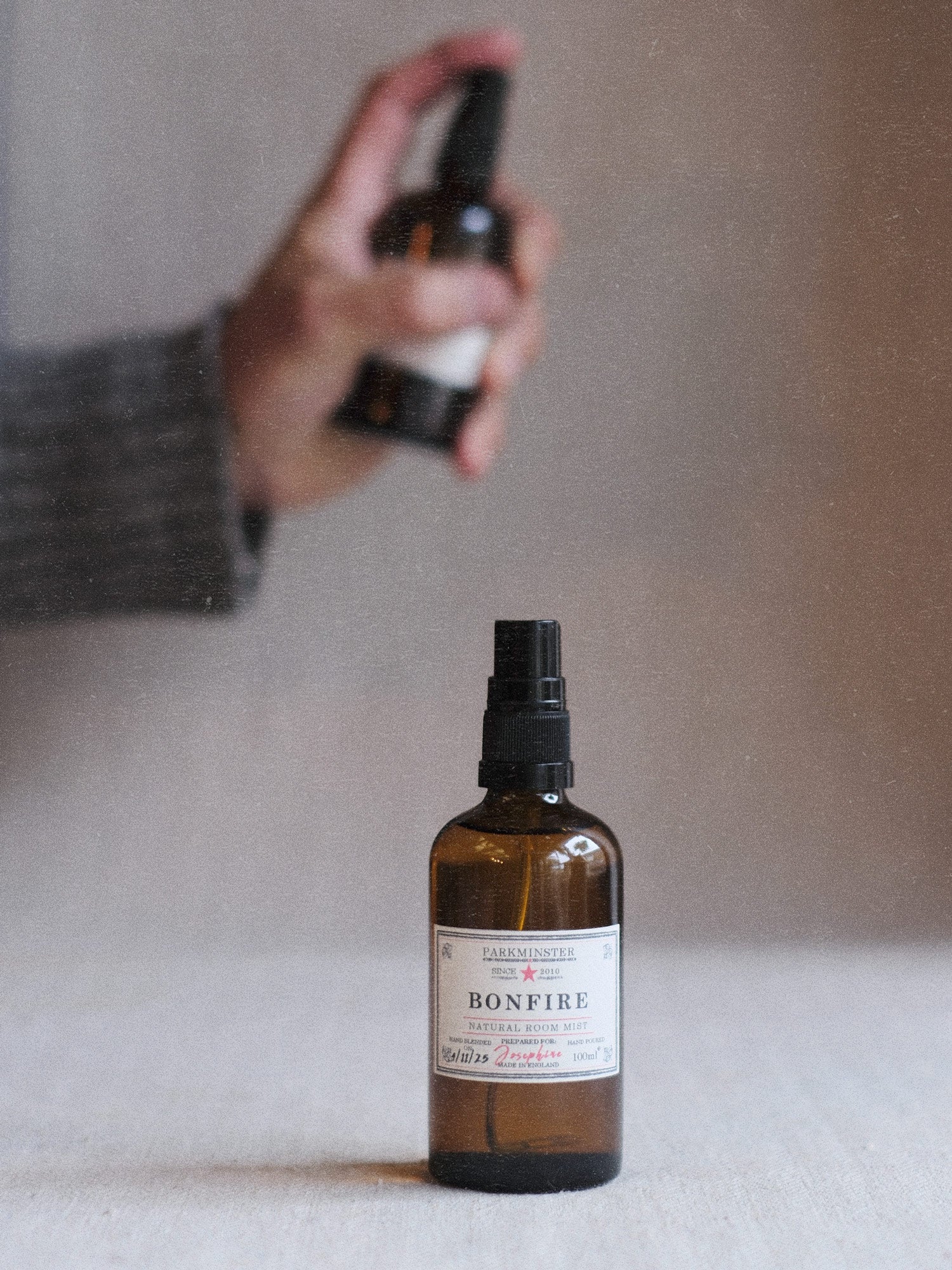 A bottle of Parkminster Bonfire Room Mist being sprayed, showing its amber glass and personalised label. A plant-based, smoky, and warm home fragrance handmade in England.