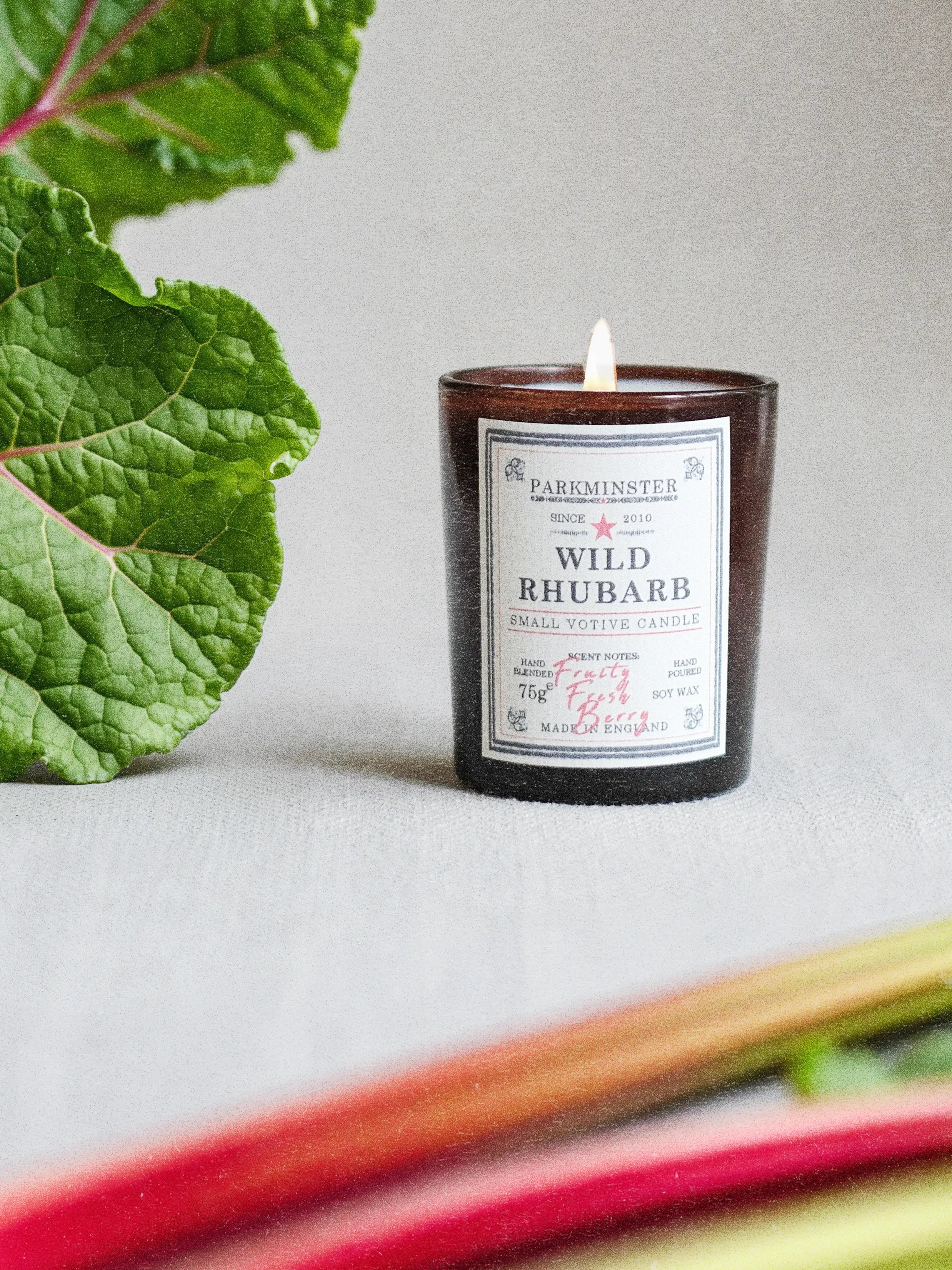 a Parkminster Wild Rhubarb 75g natural soy wax votive candle in amber glass with fresh rhubarb leaves, and a green apple and red berry fragrance.